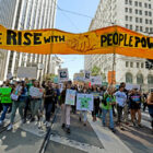 Marchers carry a banner displaying the words "We Rise With People Power"