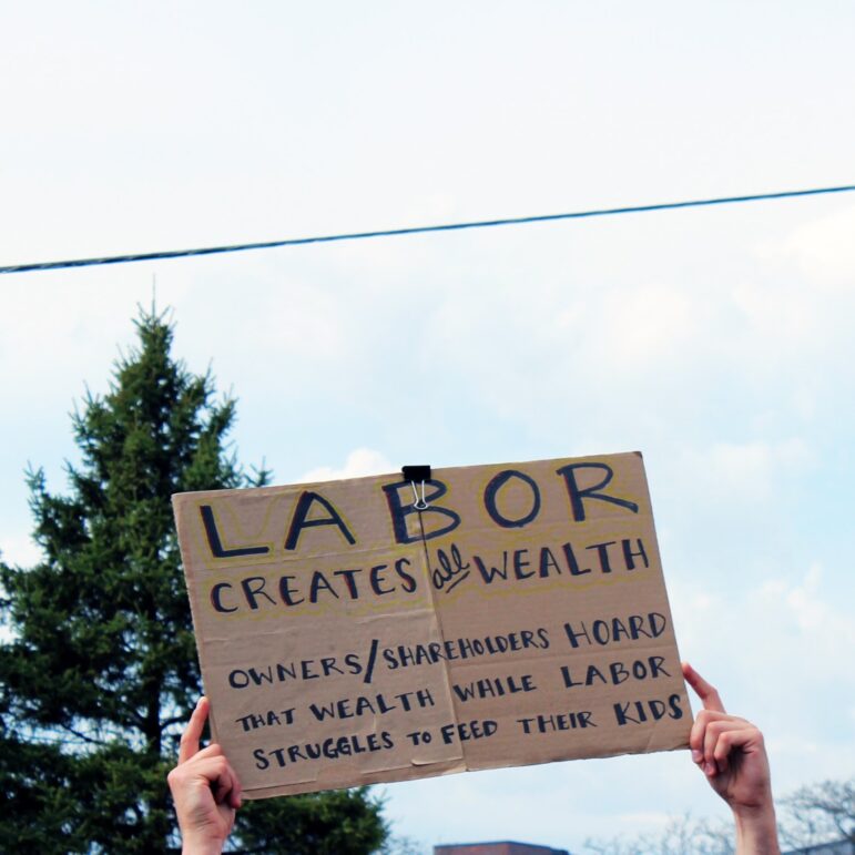 a brown cardboard sign is held up containing words in black font reading "labor creates all wealth owners/shareholders hoard that wealth while labor struggles to feed their kids"