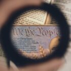a blurry hand holds up a lens to the part of the US constitution that reads "we the people"