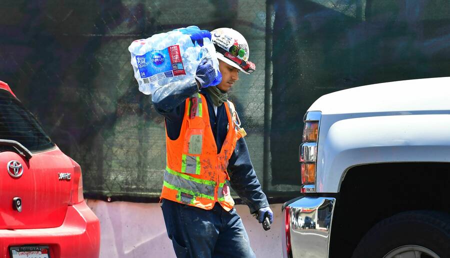 A construction worker in a neon orange vest and white hard hat with stickers carries a bundle of plastic water bottles between two cars