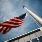 a bottom up view of an american flag attached to a tall silver pole flying in the.wind in front of a grey office building with windows