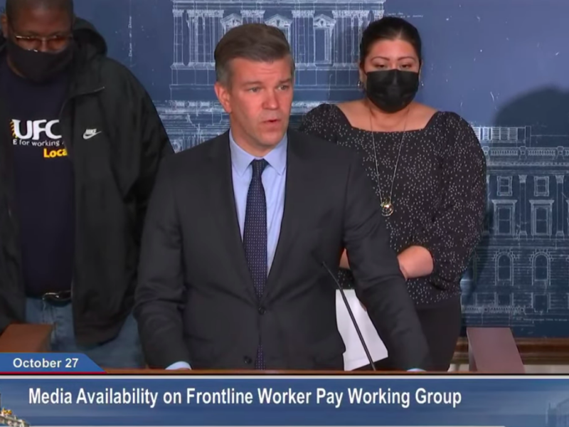 screenshot of press conference man in dark suit and blue shirt standing behind a podium in front of two people one wearing a black sweatshirt and other wearing a black dress and black face mask