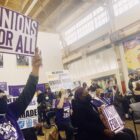 a group of workers wearing purple tee shirts hold up signs, one worker toward the front left holds up a sign saying "unions for all"