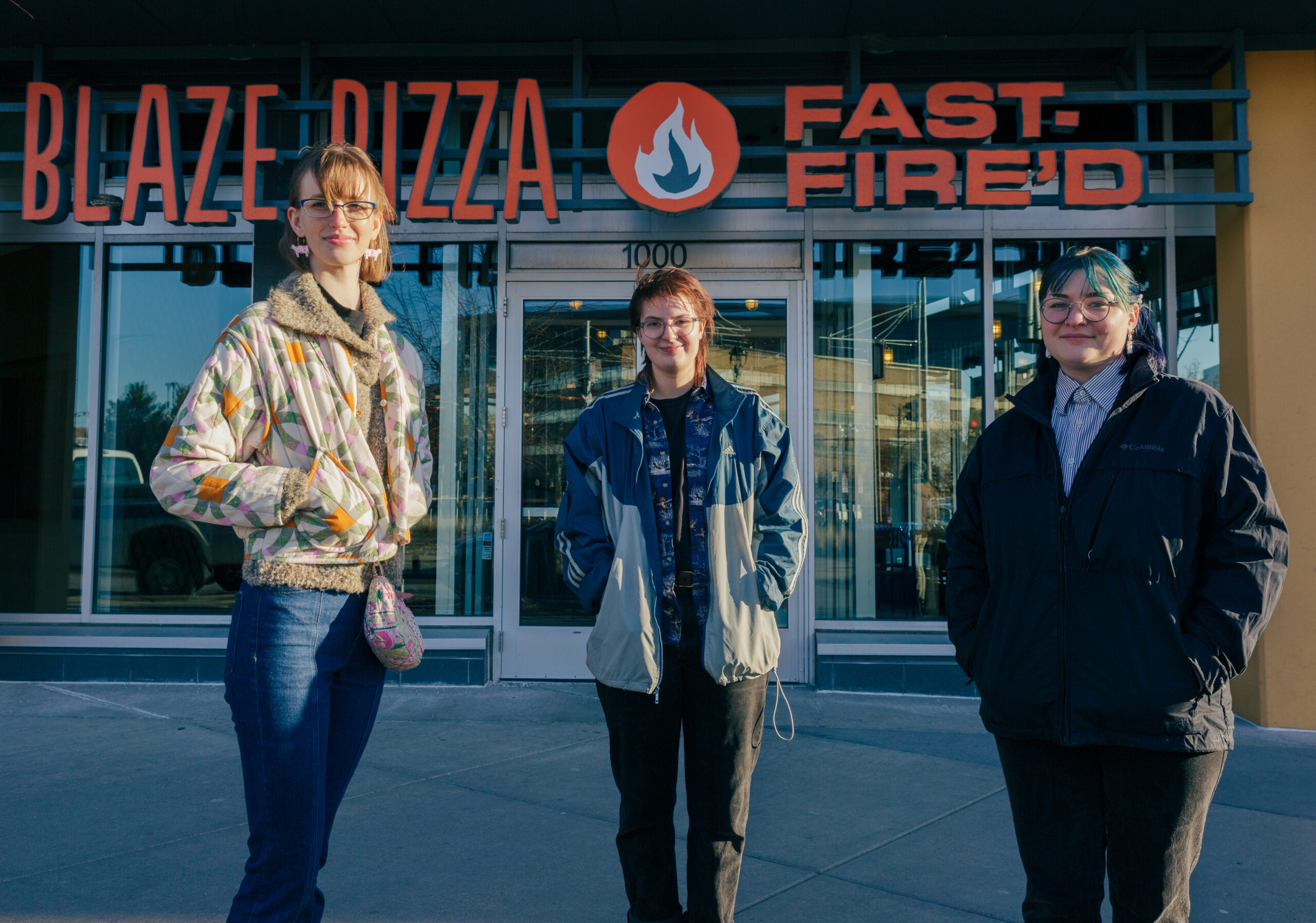 three people stand in front of the blaze pizza storefront, one wears a khaki jacket, one a denim jacket, and one a black jacket with blue hair