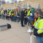 a man in a neon yellow construction jacket holds a silver bell, while people, some wearing neon yellow vests and white hard hats, line up in front of the worker's memorial with 13 white crosses