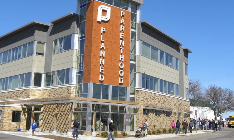 the front of the beige building with the planned parenthood name and logo, a line of people are at the bottom right of the image