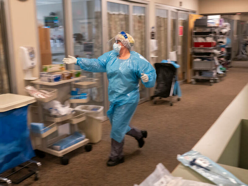 a healthcare worker wearing a blue PPE suit and white mask, gloves, and face shield rushes down a hospital hallway reaching out a hand to a hand sanitizer. the background is slightly blurred to convey movement
