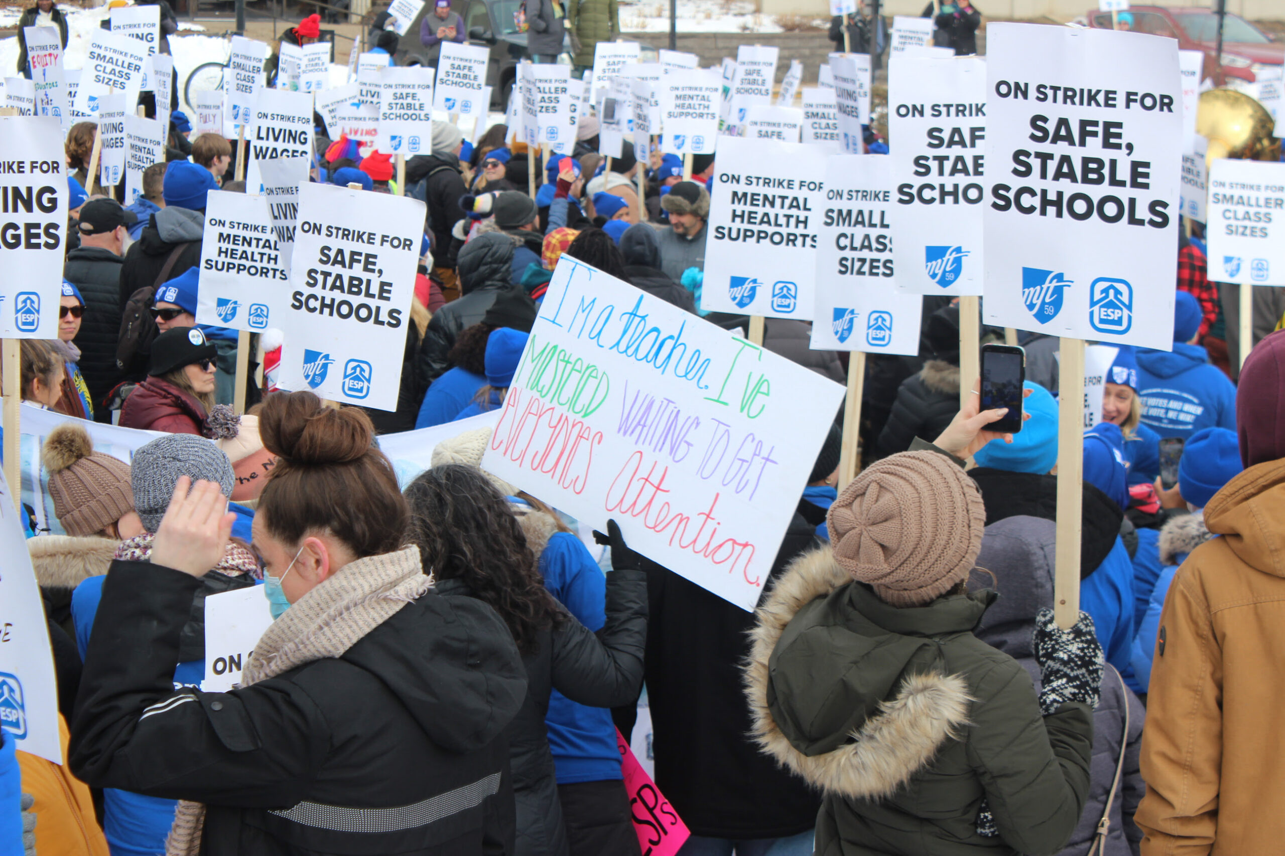 crowd of people holding signs that read "safe and stable schools" and one in the middle reading "i'm a teacher i've mastered waiting to get people's attention"