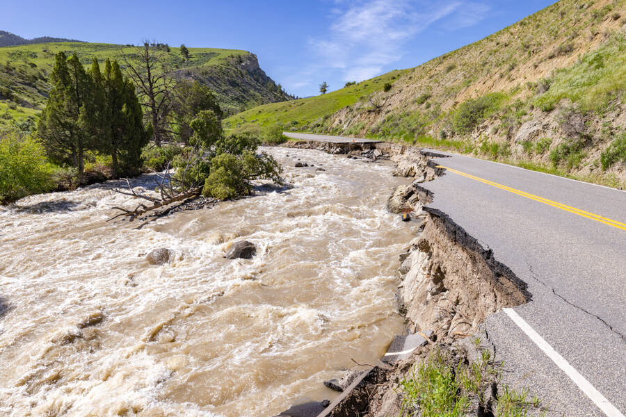 an asphalt road that cuts through green hills is eroded by brown floodwater