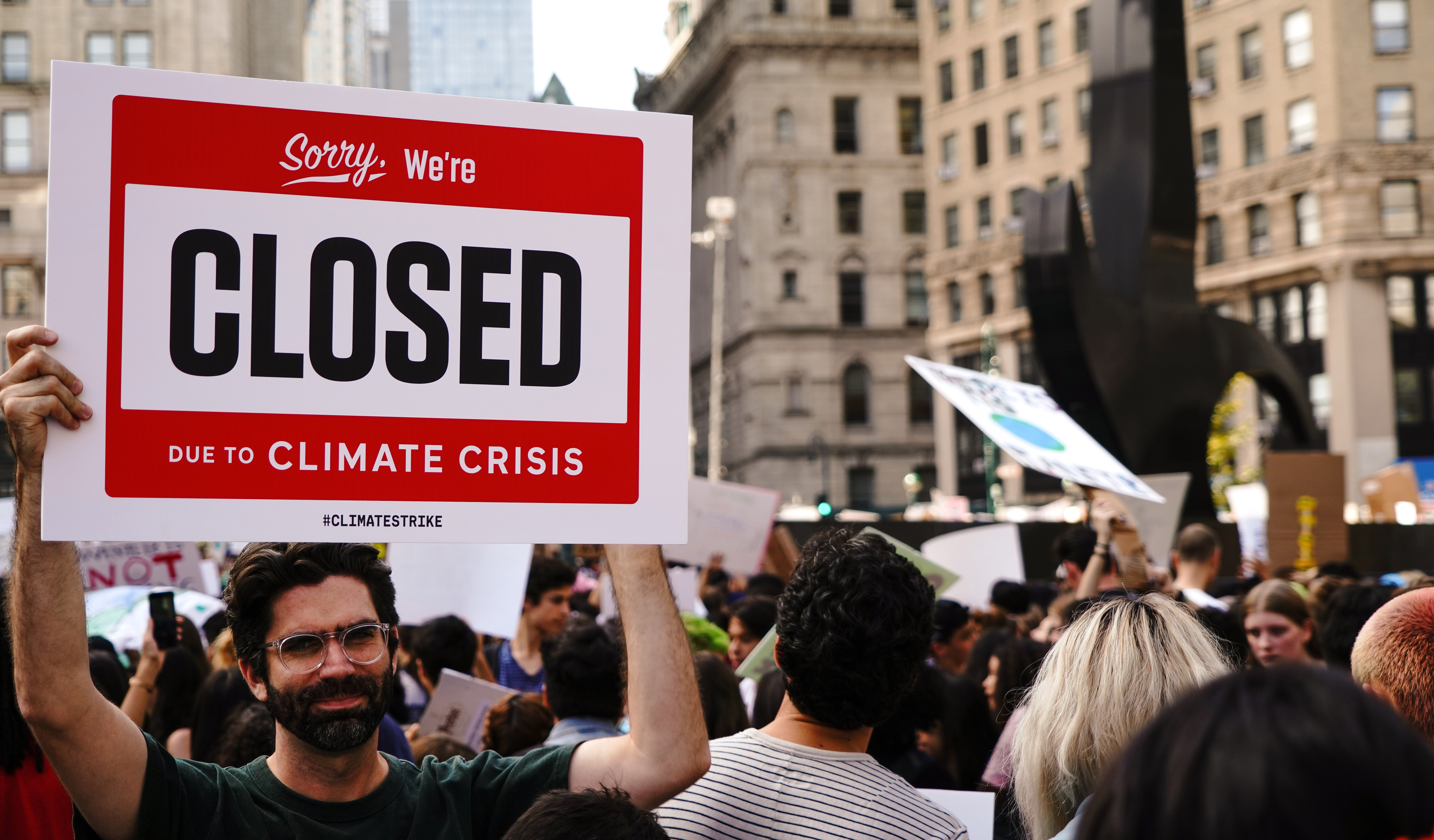a young man in a crowd holds up a sign reading "sorry we're closed due to the climate crisis"