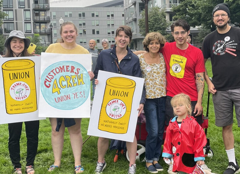 7 adults and one child stand in a row smiling holding blue, yellow, and white signs depicting a trader joe's soup can that says "union"