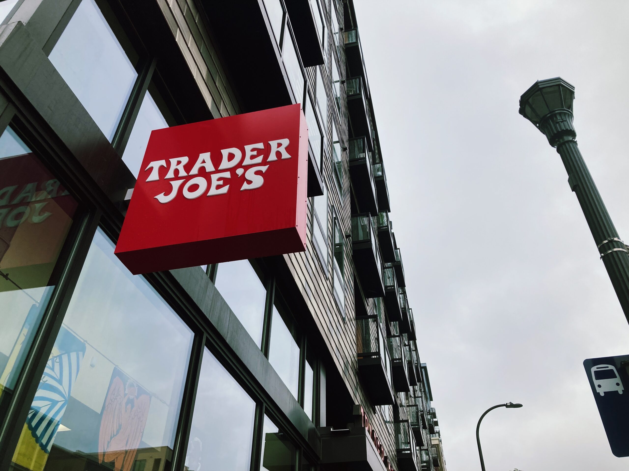 looking up at a red shop sign that reads "trader joe's" in white, a lampost and building are in the background