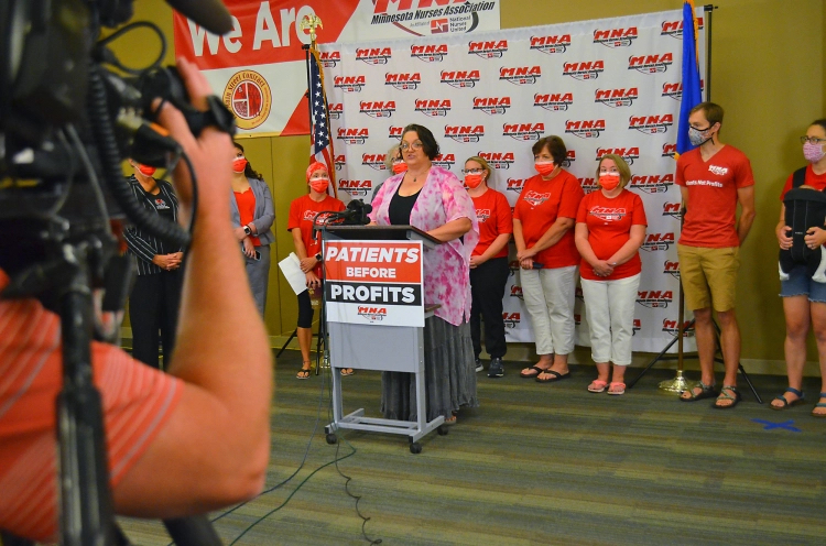 a woman in a pink and white blouse stands behind a podium with a sign on it that reads "patients before profits" in red, white, and black. a camera in the foreground is pointed at her and a line of nurses in red shirts lined up behind her in front of a backdrop with the MNA logo