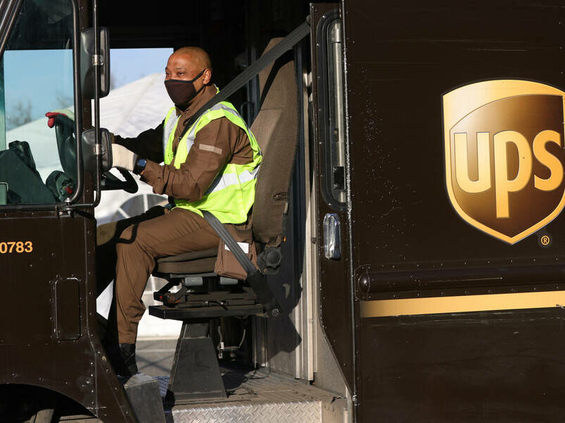A Black man wearing a black mask and a neon yellow vest sits in the driver's seat of a brown UPS truck with his hands on the wheel. The brown and yellow UPS logo is on the right side of the image of the truck