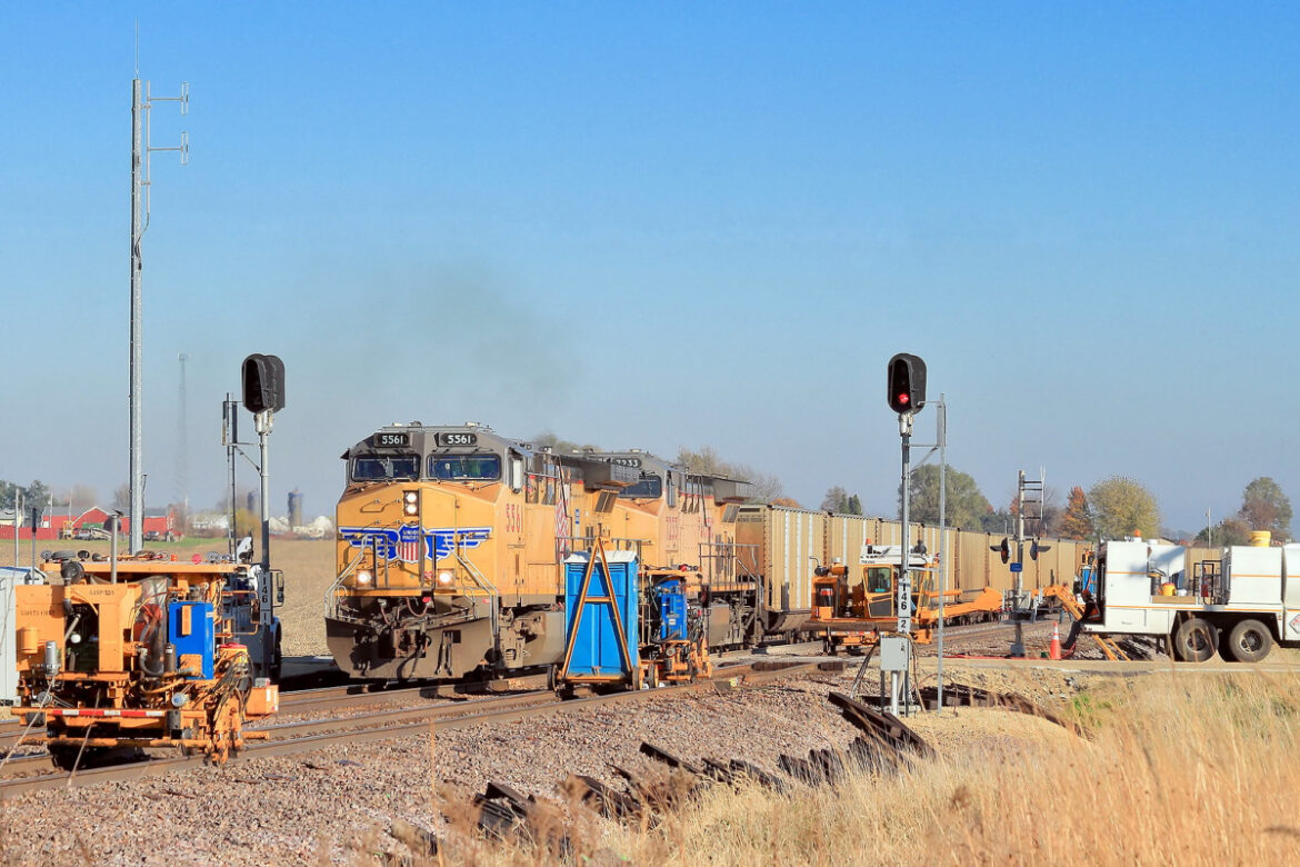 A train hauling beige containers is stopped at a red light while a small cloud of dust appears.