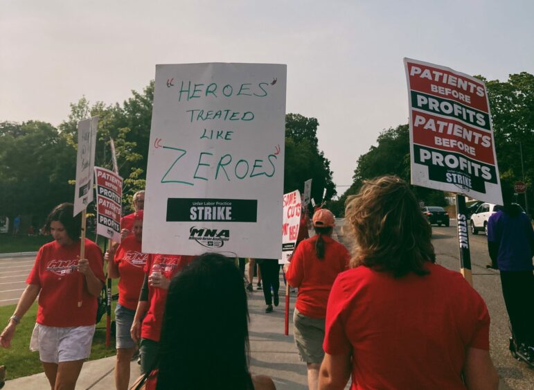 Protestors wearing red shirts move up and down the picket line, two with their backs to the camera. One sign reads ""heroes" treated like "zeroes"" and another "patients before profits"