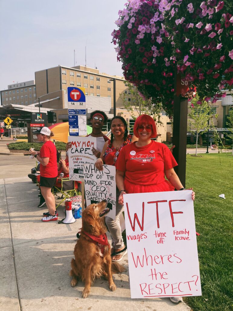 Three smiling protestors wearing red shirts and one with a red wig hold their signs as a golden retriever looks up to one of the humans. the sign in the front reads "WTF: wages, time off, family leave. Where's the respect?"