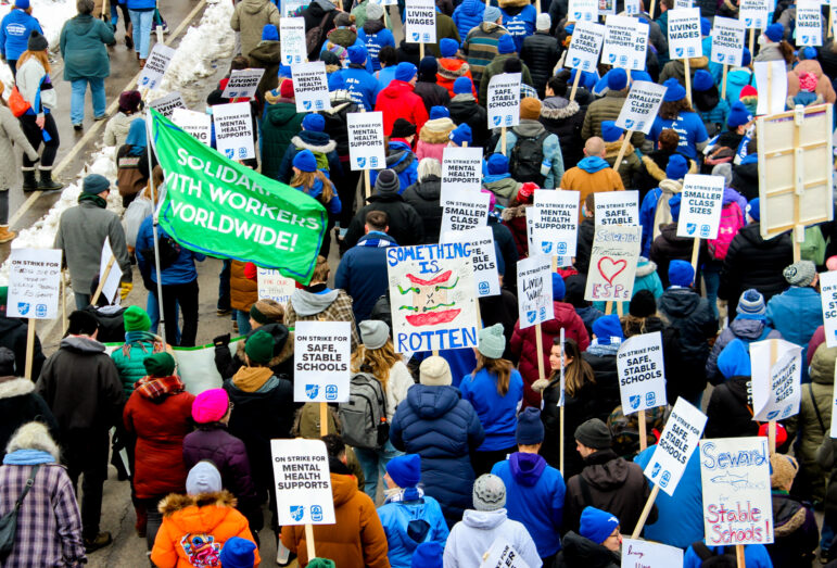 An aerial view of a crowd of protestors walking fills the image. They all hold signs, one holds a large green flag with white text reading "solidarity with workers worldwide!" and another sign depicts a red apple with green worms coming out of it reading "something is rotten"