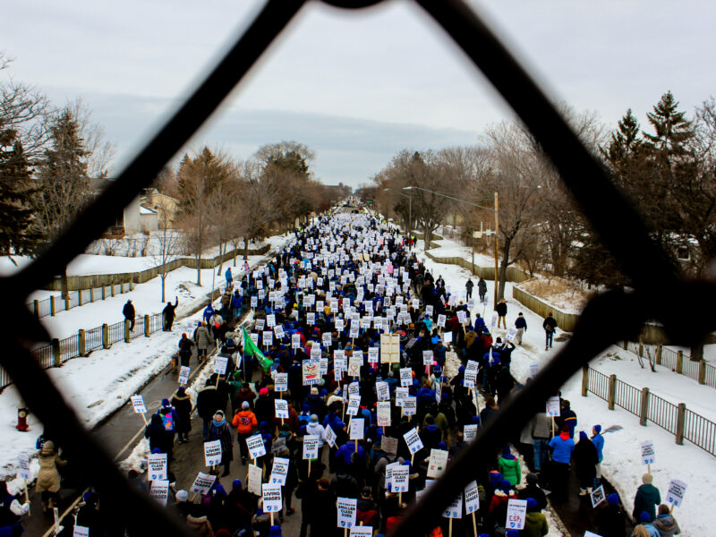 an aerial view of a protest march in the snow-lined streets, framed by a chain-link fence.