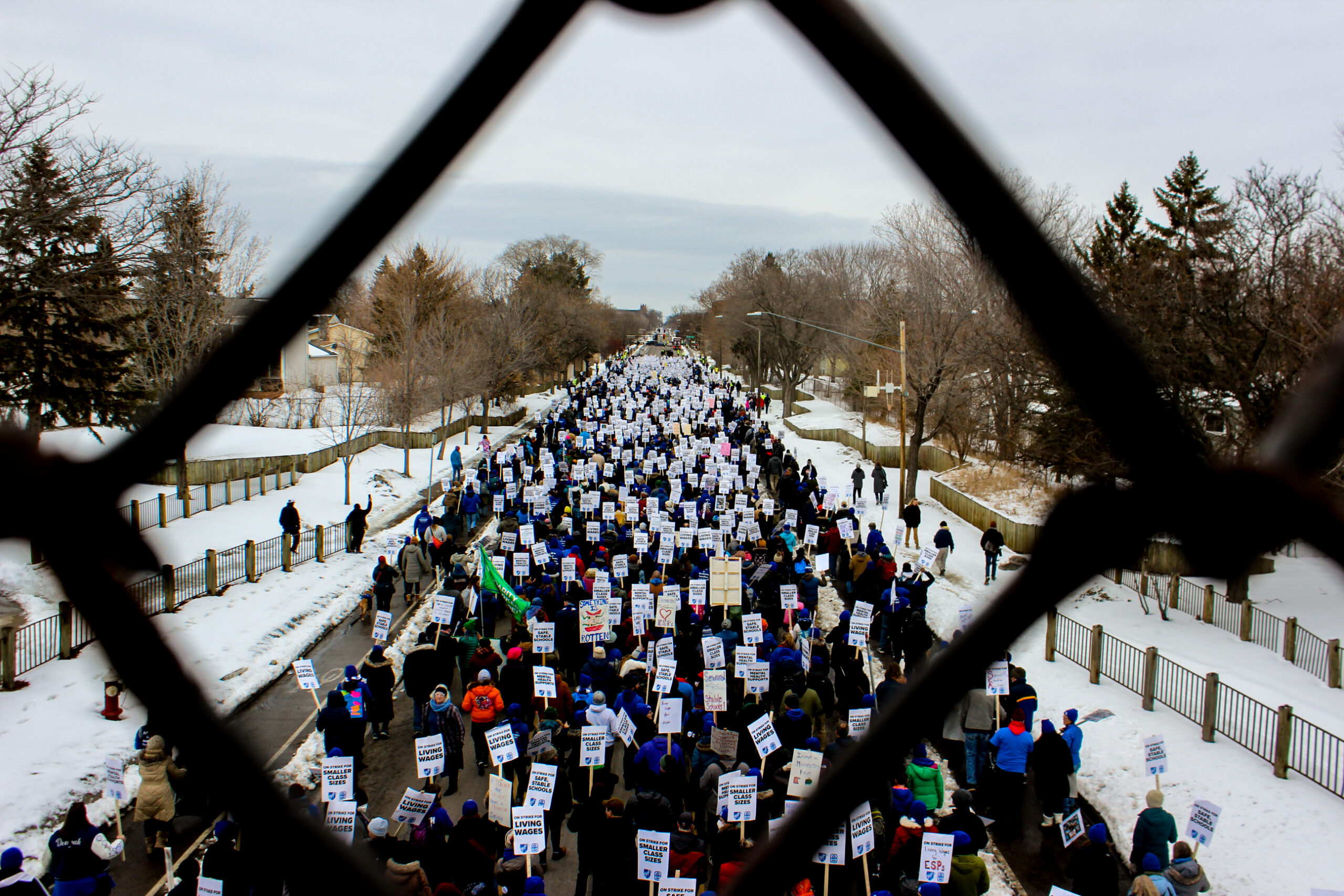 an aerial view of a protest march in the snow-lined streets, framed by a chain-link fence.