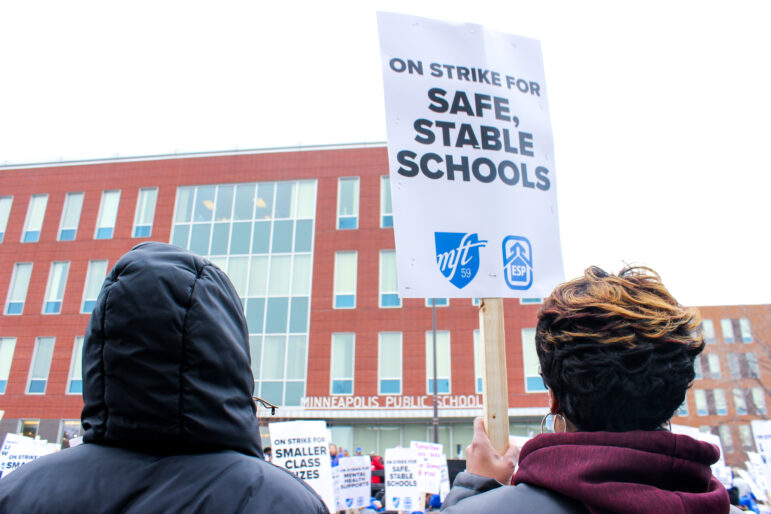 two Black women stand with their backs to the camera looking at the office building of Minneapolis Public Schools District. One holds a sign reading "on strike for safe, stable schools" with the MFT 59 and ESP chapter logos
