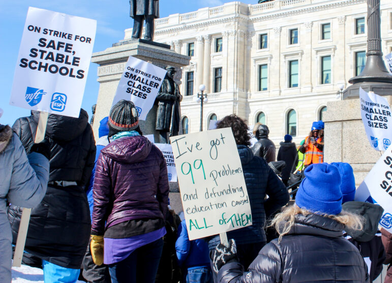 A handful of people standing in a crowd hold picket signs in front of the white state Capitol building. The sign in the middle reads "I've got 99 problems and defunding education caused all of them."