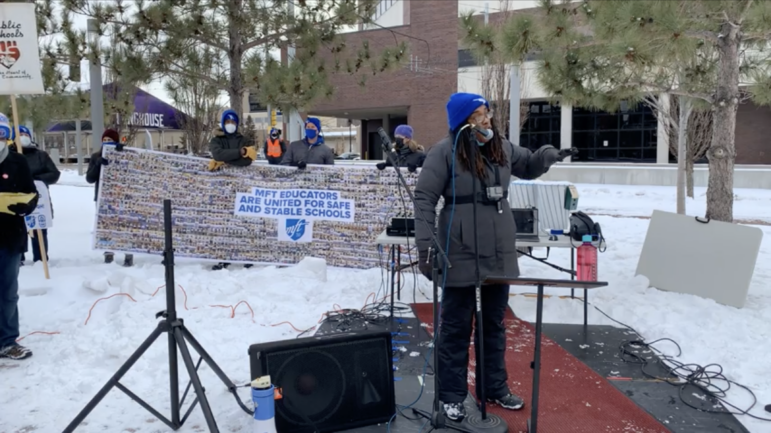 A screenshot of a livestream showing a Black woman wearing a black coat and blue hat and go pro attached to her chest standing behind a microphone in front of an MFT 59 banner