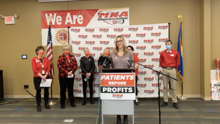 Screenshot of a livestream where a blonde woman stands behind a podium, in front of others wearing red shirts and a large white banner with the red MNA logo pattern
