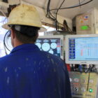 a man in blue jumpsuit and white hard hat stands in front of two monitors and a window at an oil rig