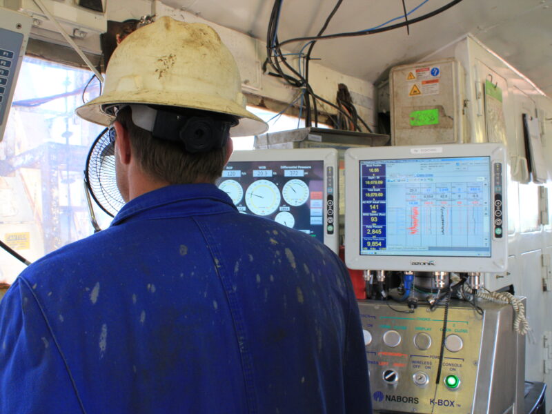 a man in blue jumpsuit and white hard hat stands in front of two monitors and a window at an oil rig