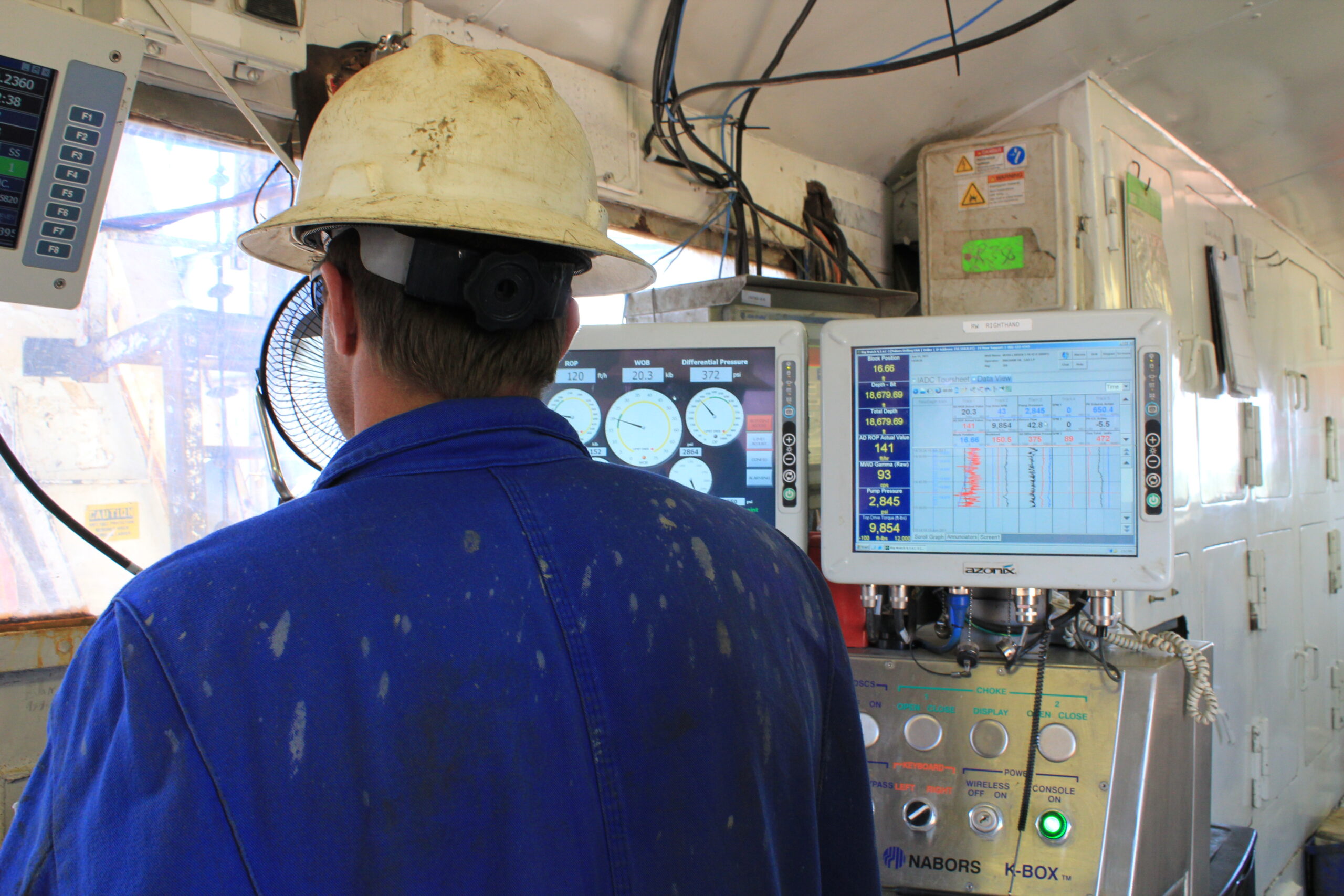 a man in blue jumpsuit and white hard hat stands in front of two monitors and a window at an oil rig