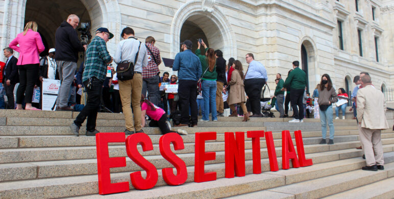 red letters spelling out the word "essential" line the steps of the state capitol building in front of legs of a crowd