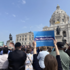 The backs of people standing in a crowd with the white marble state capitol building in the background against a blue sky with scattered white clouds. A brunette woman holds up a blue sign reading "We're Making Minnesota the #BestState for Families, Children, and Workers!"