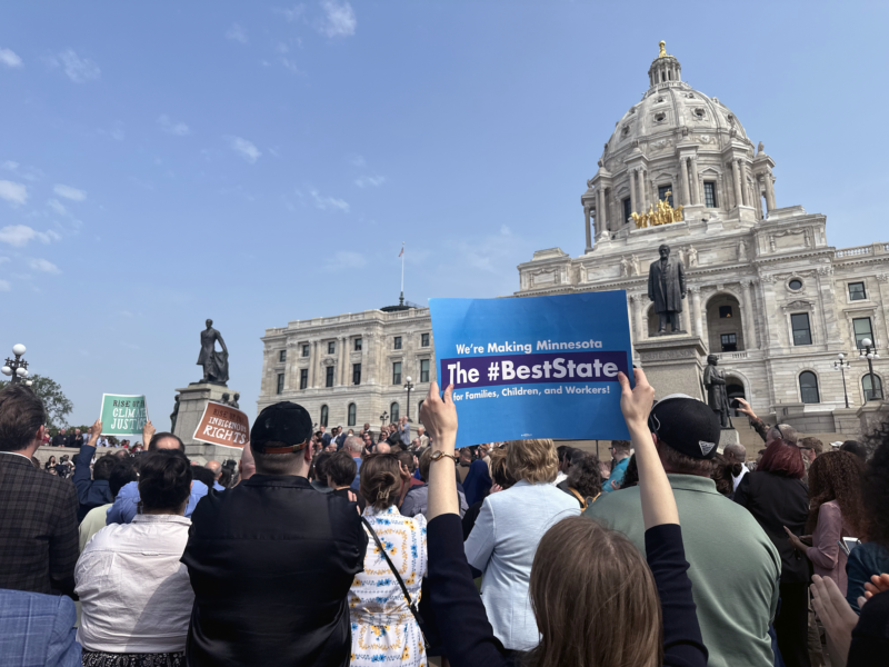The backs of people standing in a crowd with the white marble state capitol building in the background against a blue sky with scattered white clouds. A brunette woman holds up a blue sign reading "We're Making Minnesota the #BestState for Families, Children, and Workers!"