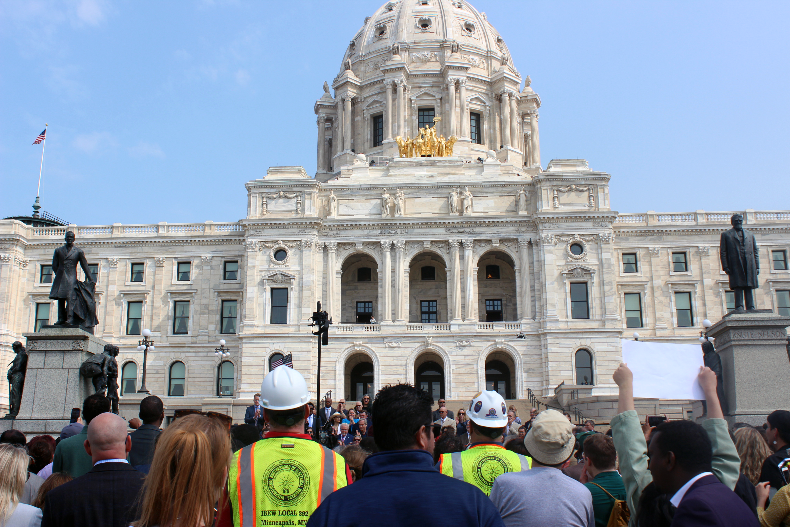 A crowd of people stand at the foot of the white marble state capitol building steps. Two members in the crowd wear white construction helmets and neon yellow and orange vests reading IBEW Local 222, Minneapolis MN. One member of the crowd holds up a sign that is blank on the back.