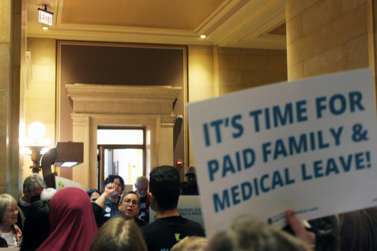 a crowd of people line the hallways of the capitol building, the face of a woman peeks out of the crowd in the middle of speaking, a sign to the right of the image reads "It's time for paid family & medical leave!"