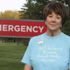 A brown haired woman stands in front of a large red emergency sign on a grass lawn wearing a sky blue tee shirt reading "well behaved women rarely make history"