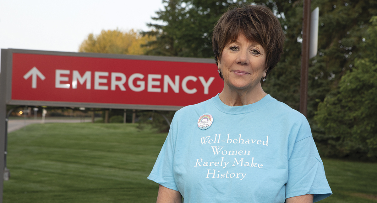 A brown haired woman stands in front of a large red emergency sign on a grass lawn wearing a sky blue tee shirt reading "well behaved women rarely make history"
