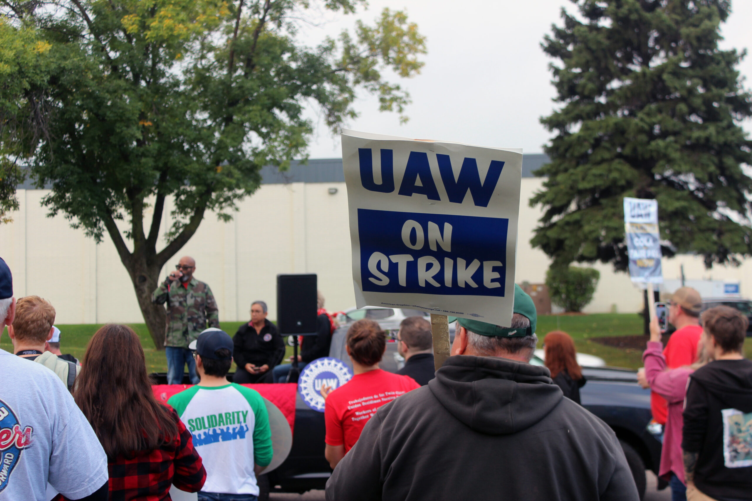 A crowd looks on to a Black man wearing a camo jacket standing next to speaker with a microphone, one crowd member holds a picket sign reading "UAW ON STRIKE"