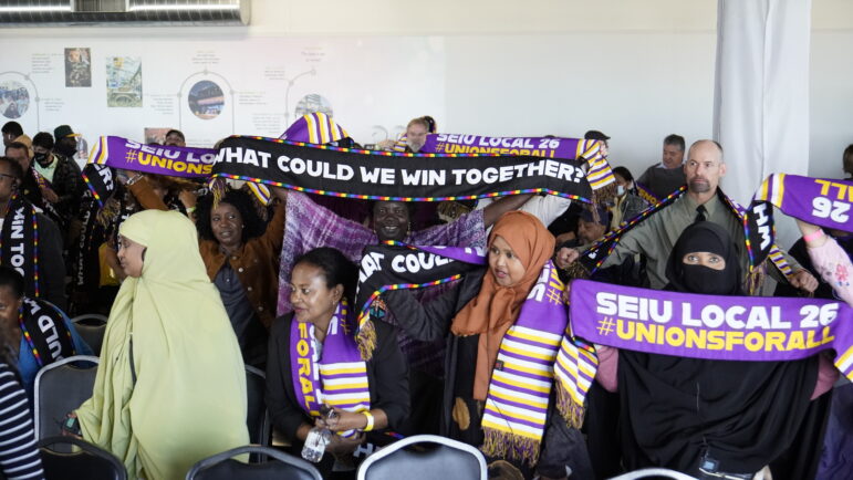 A crowd of workers hold up scarves that read "What could we win together?". 