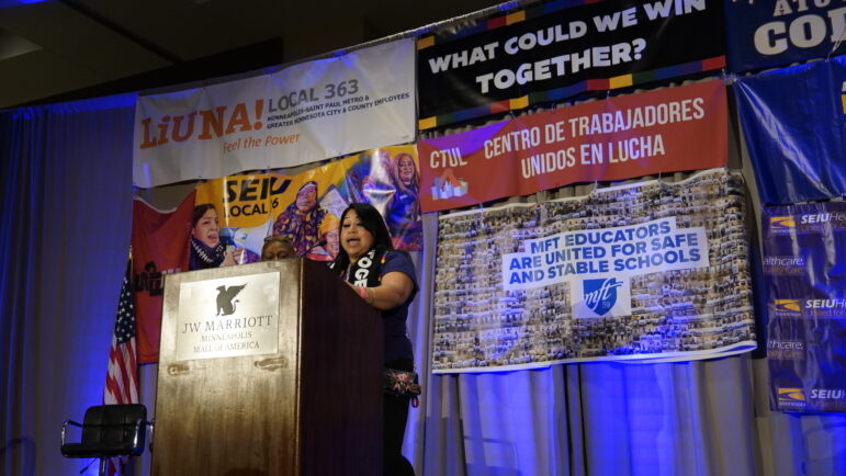 Eva Lopez on stage speaking in front of banners from all the unions and community organizations 