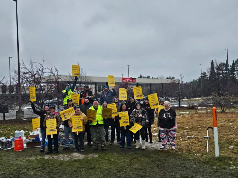 A crowd of people wearing coats and hats stand in front of a gas station holding yellow picket signs that read "Respect Us Protect Us Pay Us"