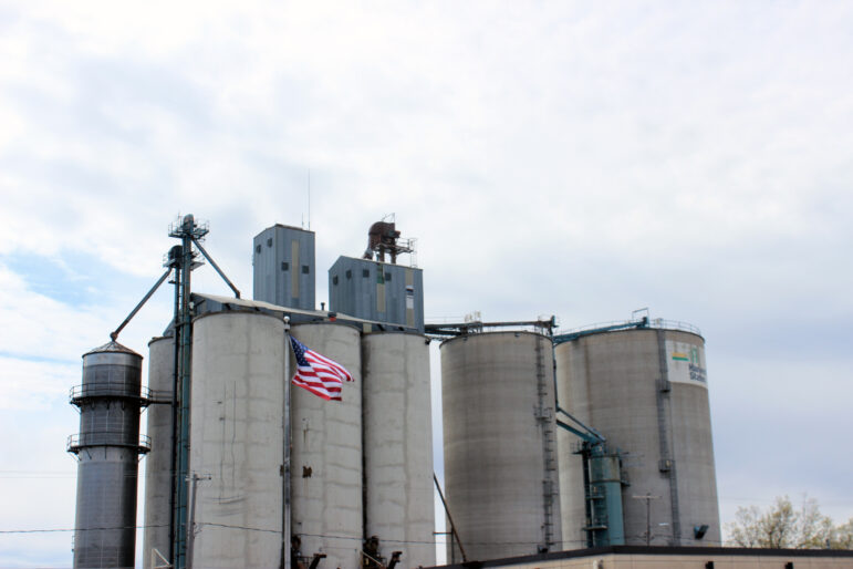 An American flag flies in the wind in front of 5 gray grain elevator silos