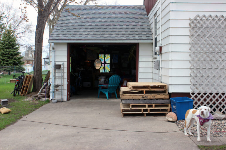 A white pitbull with brown spots on its back and legs wearing a purple bandana stands to the side of an open garage door next to wooden pallets and a blue lawn chair