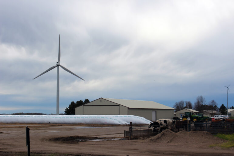 a white windmill towers over a farm site where a black and white cow stands behind fencing