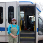 A white person with medium length curly brown hair wearing a purple bandana and blue shirt and green overalls and a grey face masks stands holding their hands in front of the open doors of a white bus with blue stripes as a bus driver smiles at the camera