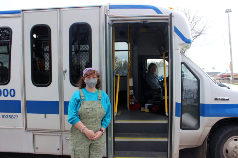 A white person with medium length curly brown hair wearing a purple bandana and blue shirt and green overalls and a grey face masks stands holding their hands in front of the open doors of a white bus with blue stripes as a bus driver smiles at the camera