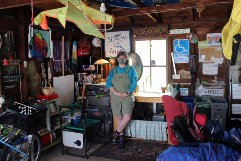 Syd stands in a garage surrounded by signs and chairs and crafts and gear