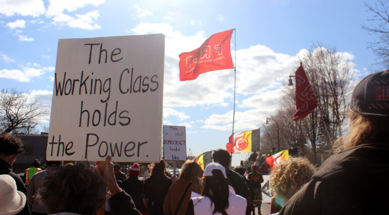 A crowd of protestors holding signs, one up close reads "The Working Class holds the Power"