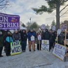 around 2 dozen people wearing winter gear, matching grey beanies, stand in the snow holding picket signs reading "on strike" and "fair wages" and "union strong SEIU"